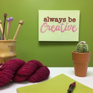 A desk with yarn and knitting needles, paper and a pen, and a sign on the wall that reads "always be creative."